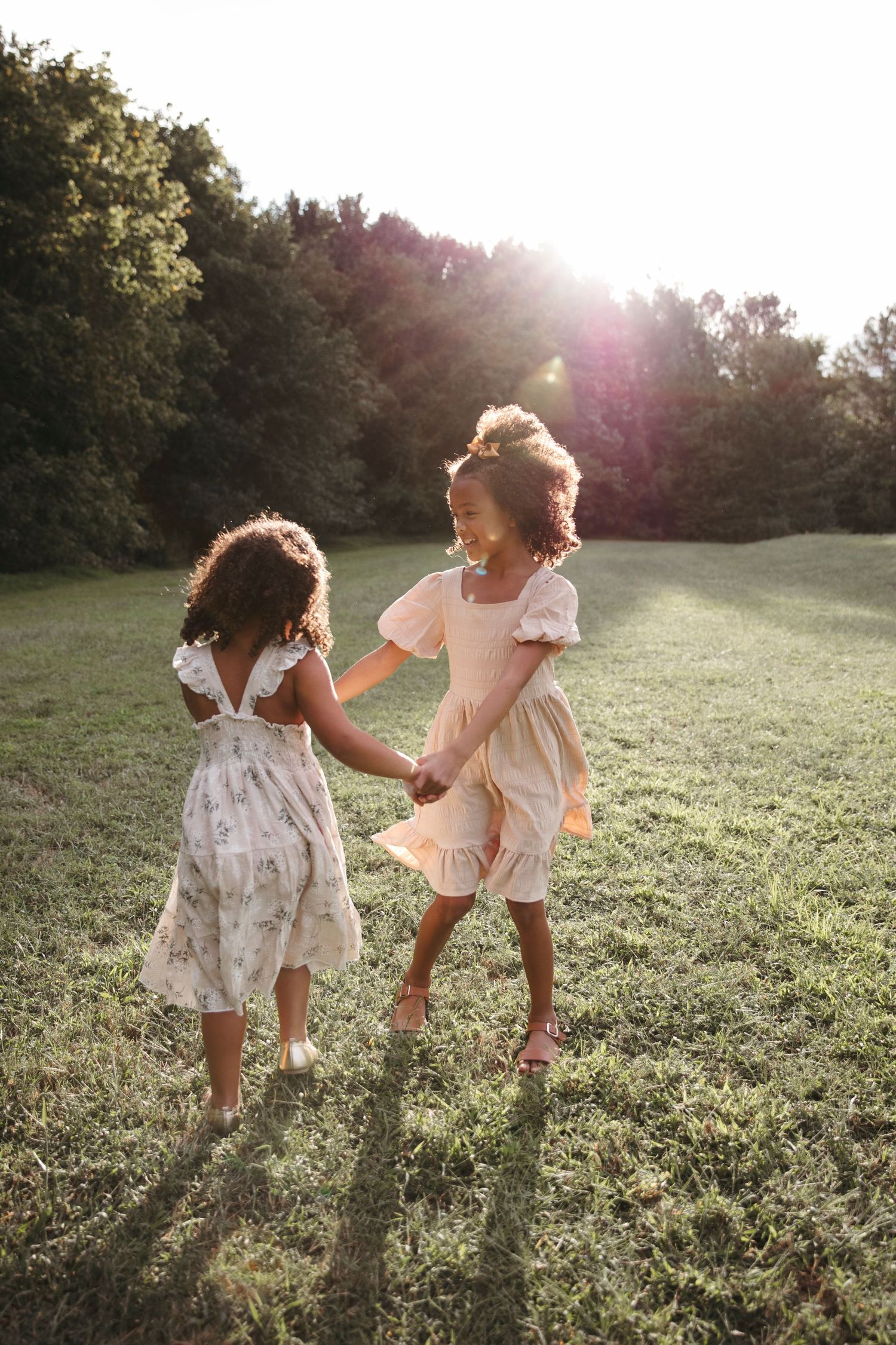 Two children in flowing dresses dance and play together in a sunny meadow at golden hour.