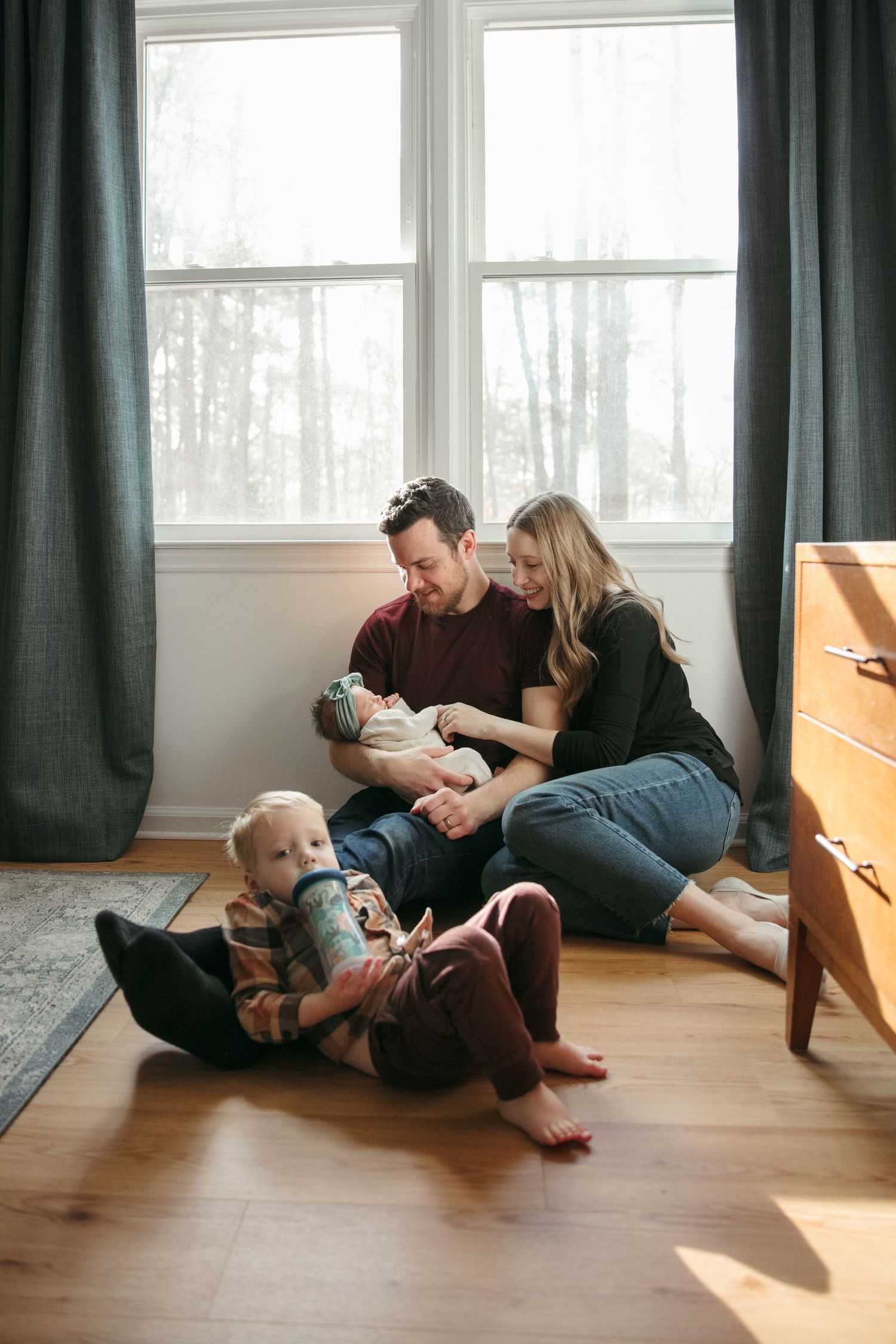 Natural light streams through windows into room with hardwood floors and dark curtains on a family with a newborn baby.