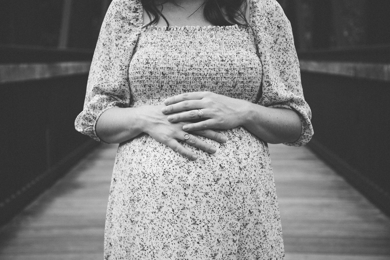 A black and white photo of a floral dress-clad figure tenderly cradling their pregnant belly on a wooden bridge.