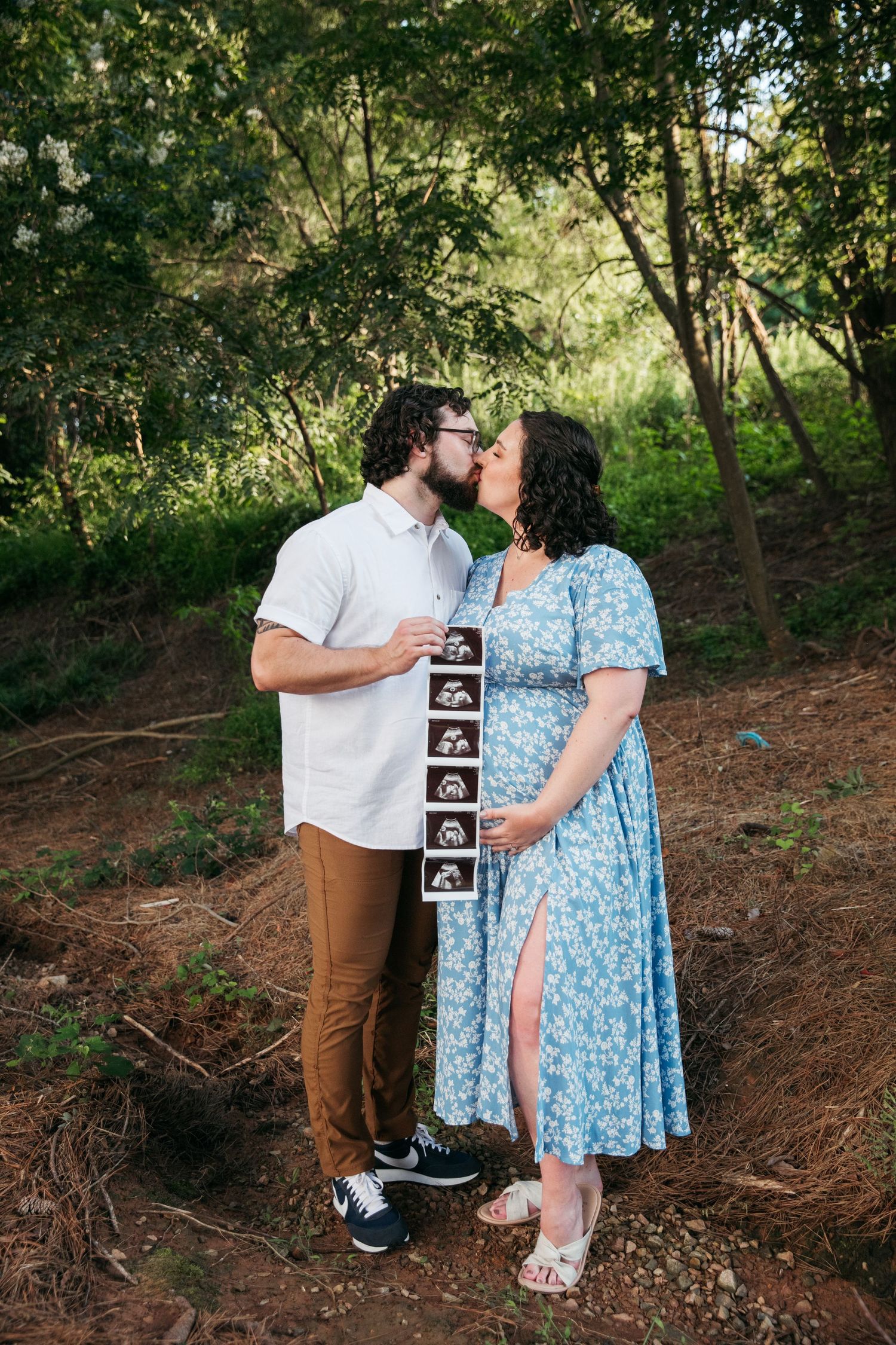 A couple holds ultrasound photos while embracing in a forest setting.