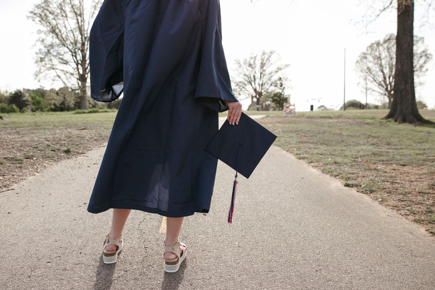 Graduate in traditional navy cap and gown walking outdoors on concrete path.