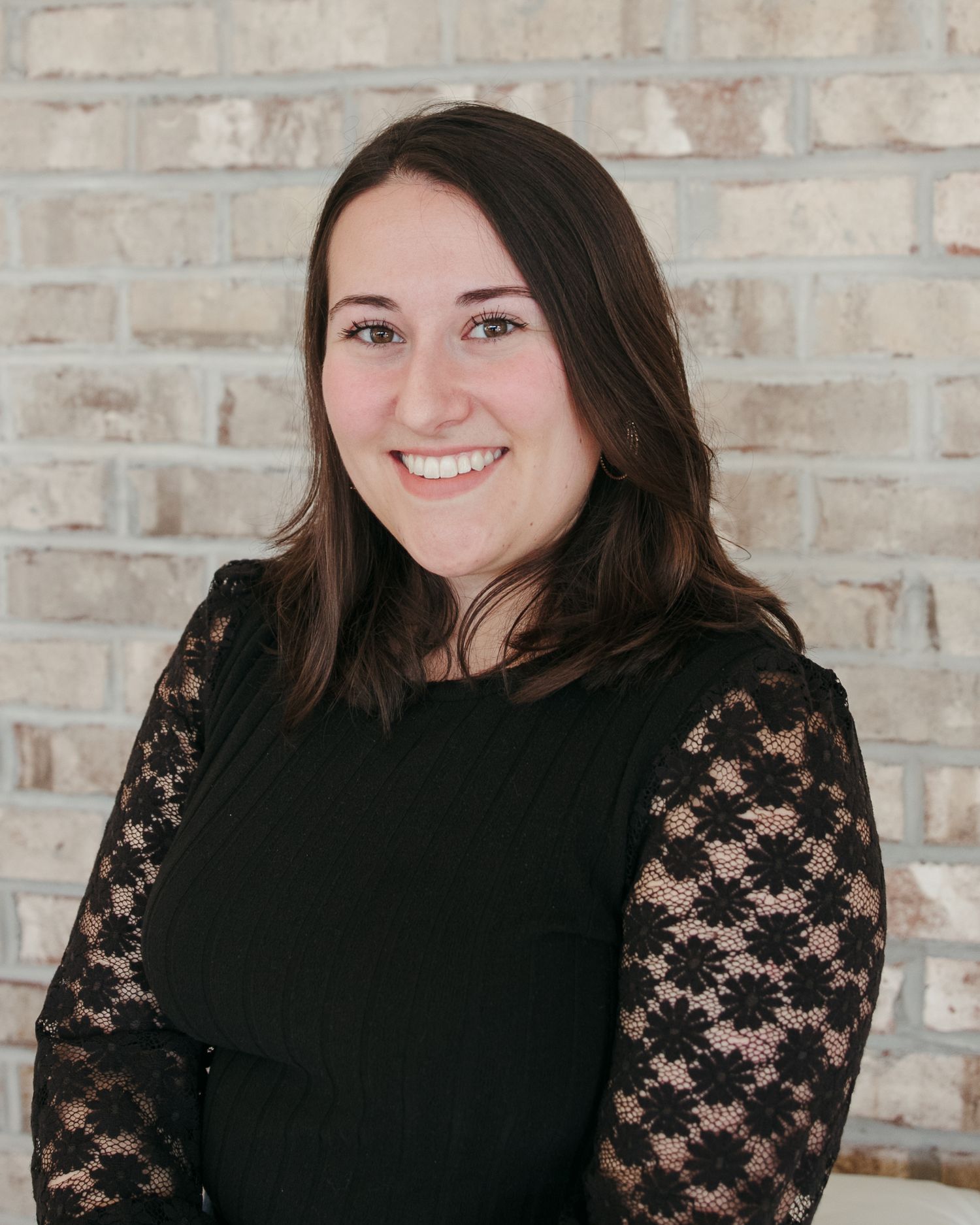 Professional headshot against brick wall wearing black top with lace sleeves.