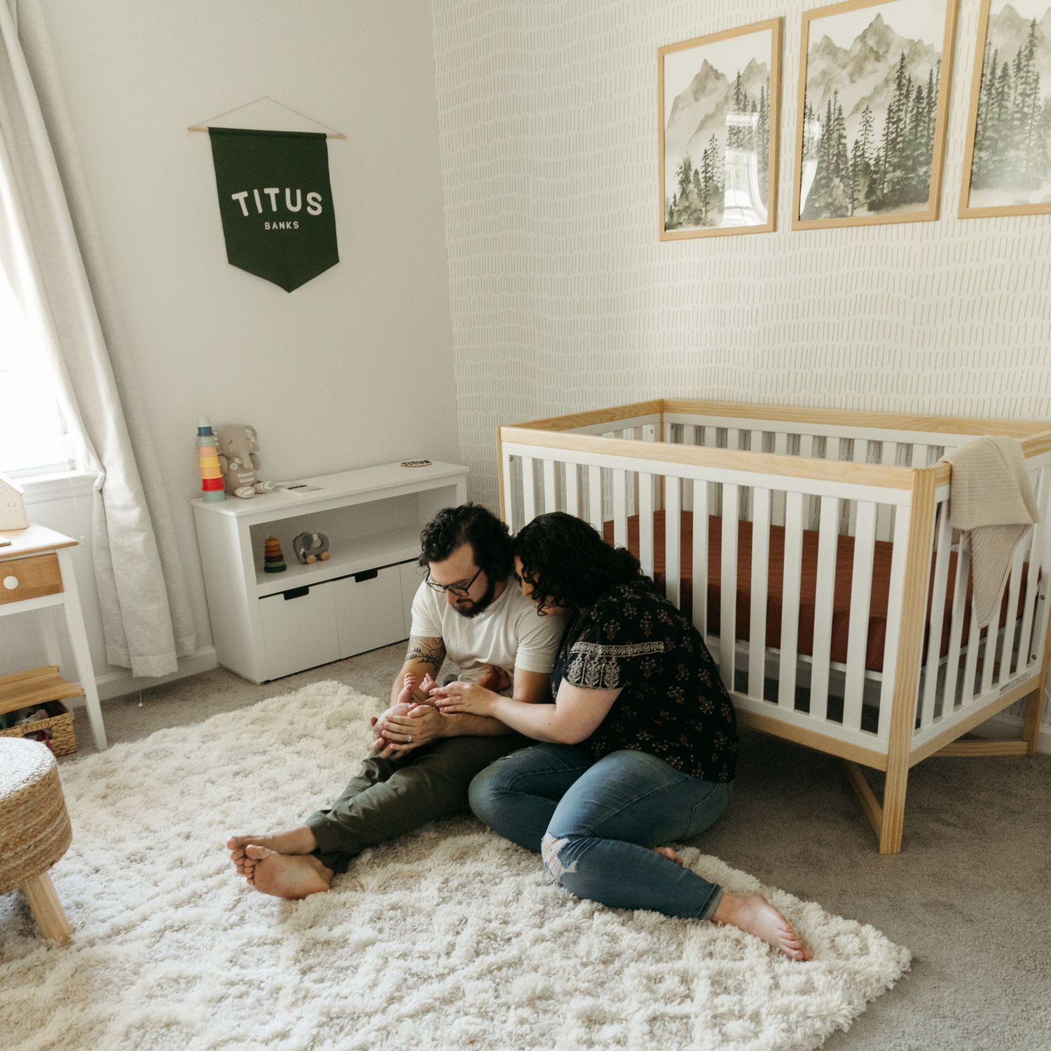 Cozy nursery scene with couple relaxing on carpet beside wooden crib under nature prints.