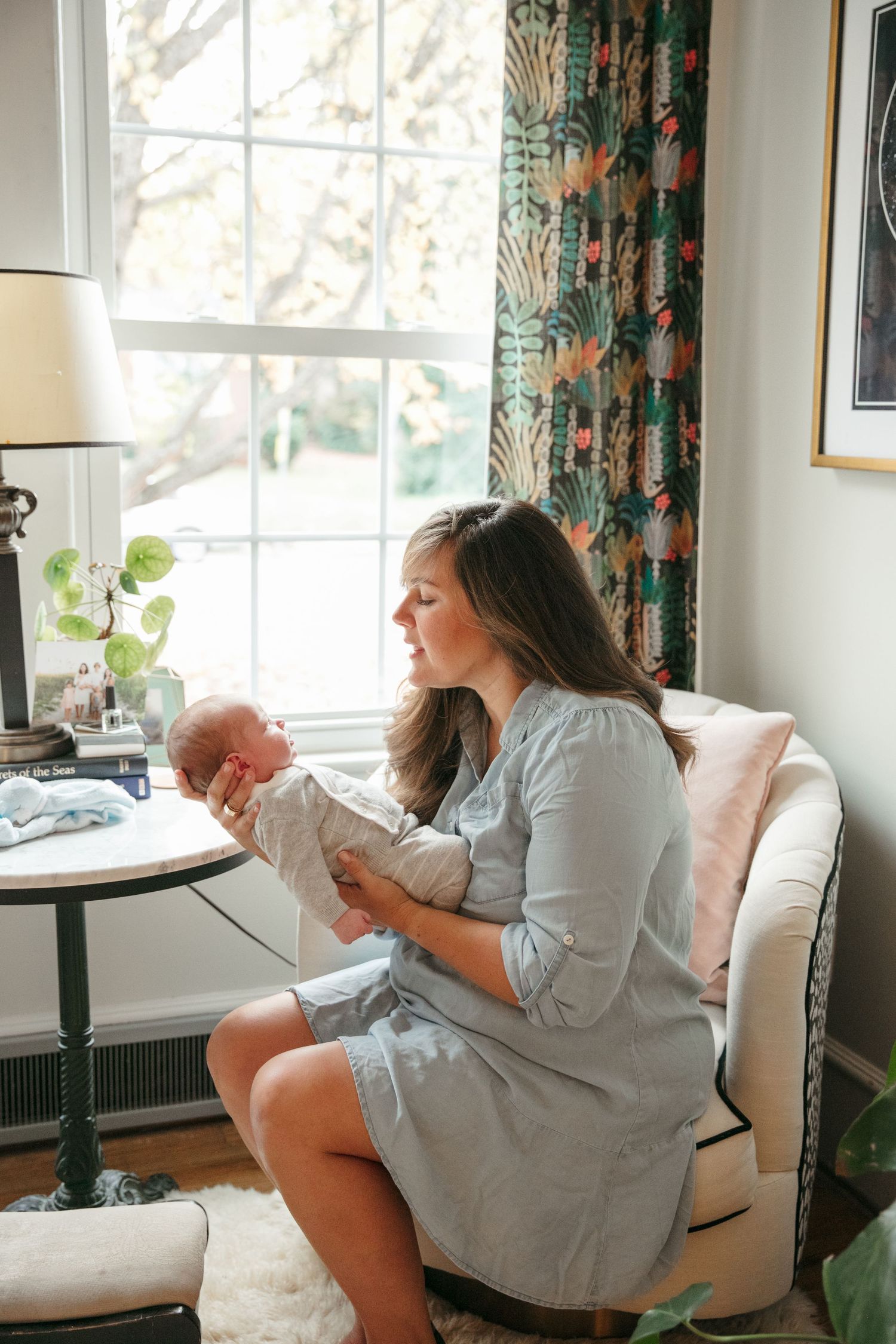 Parent in gray dress holds newborn in cozy living room with floral curtains.