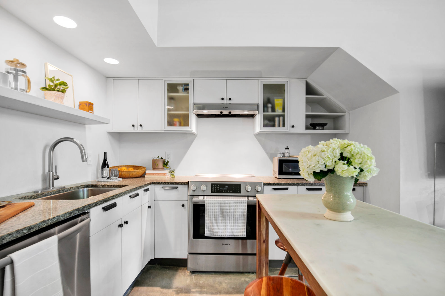 Modern white kitchen with stainless steel appliances, granite countertops, open shelving, and fresh white hydrangeas on the island.