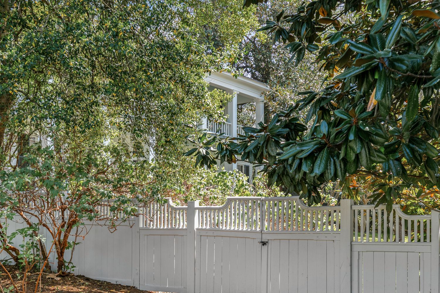 White picket fence surrounds a charming home with columns, framed by lush magnolia trees and green landscaping.