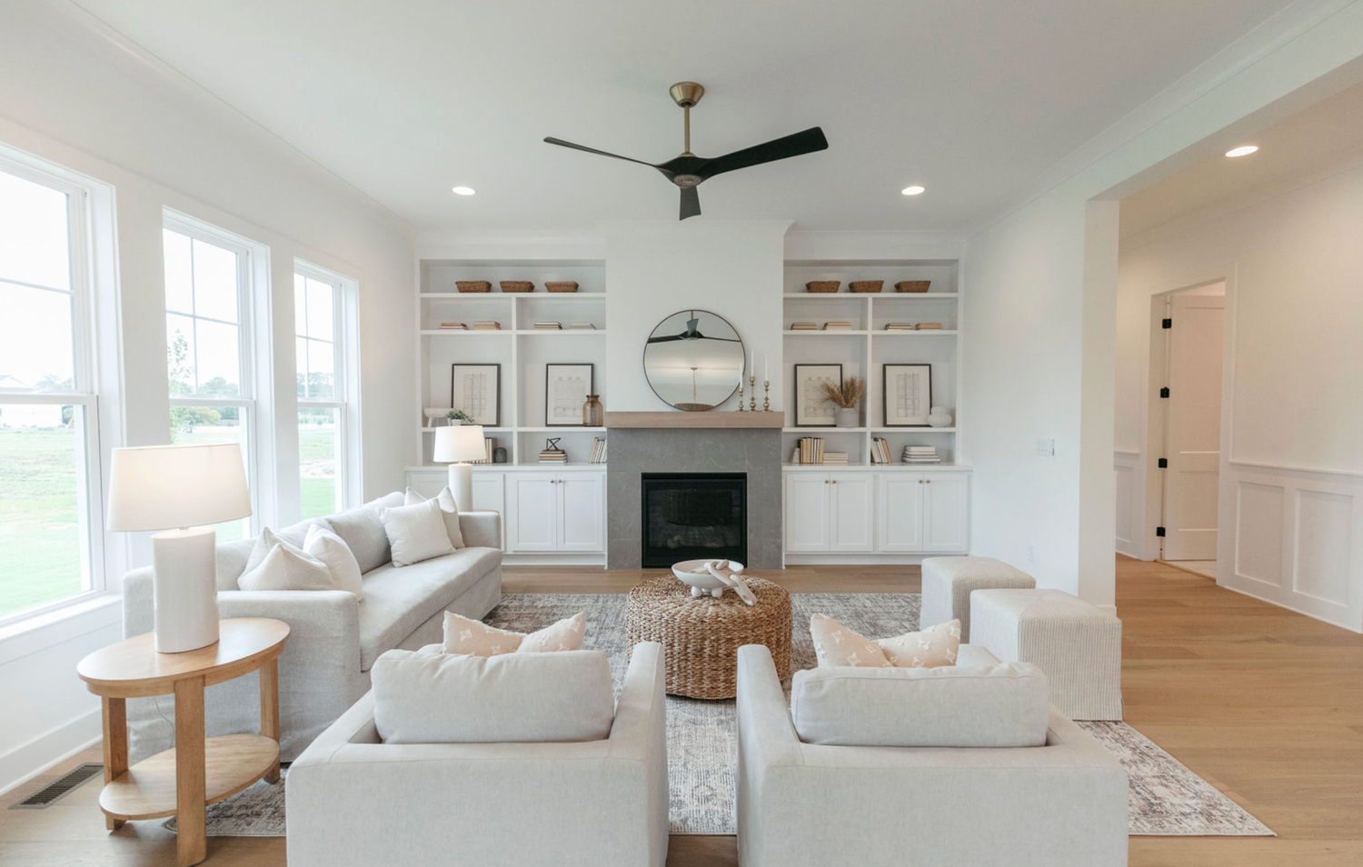 Bright white living room with built-in shelves, fireplace, ceiling fan, and neutral furniture on hardwood floors.
