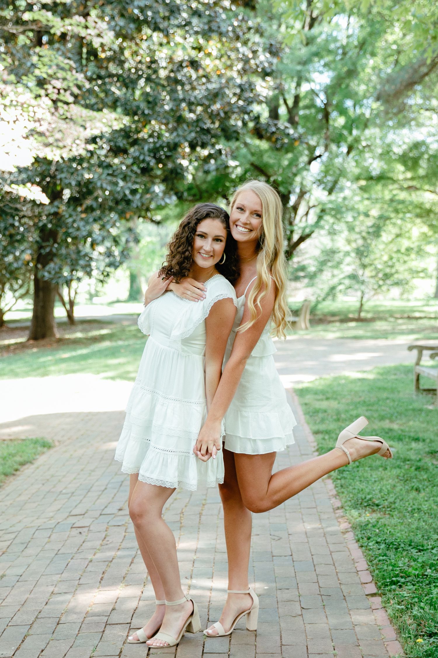 Two friends in white dresses pose playfully on brick pathway surrounded by greenery.
