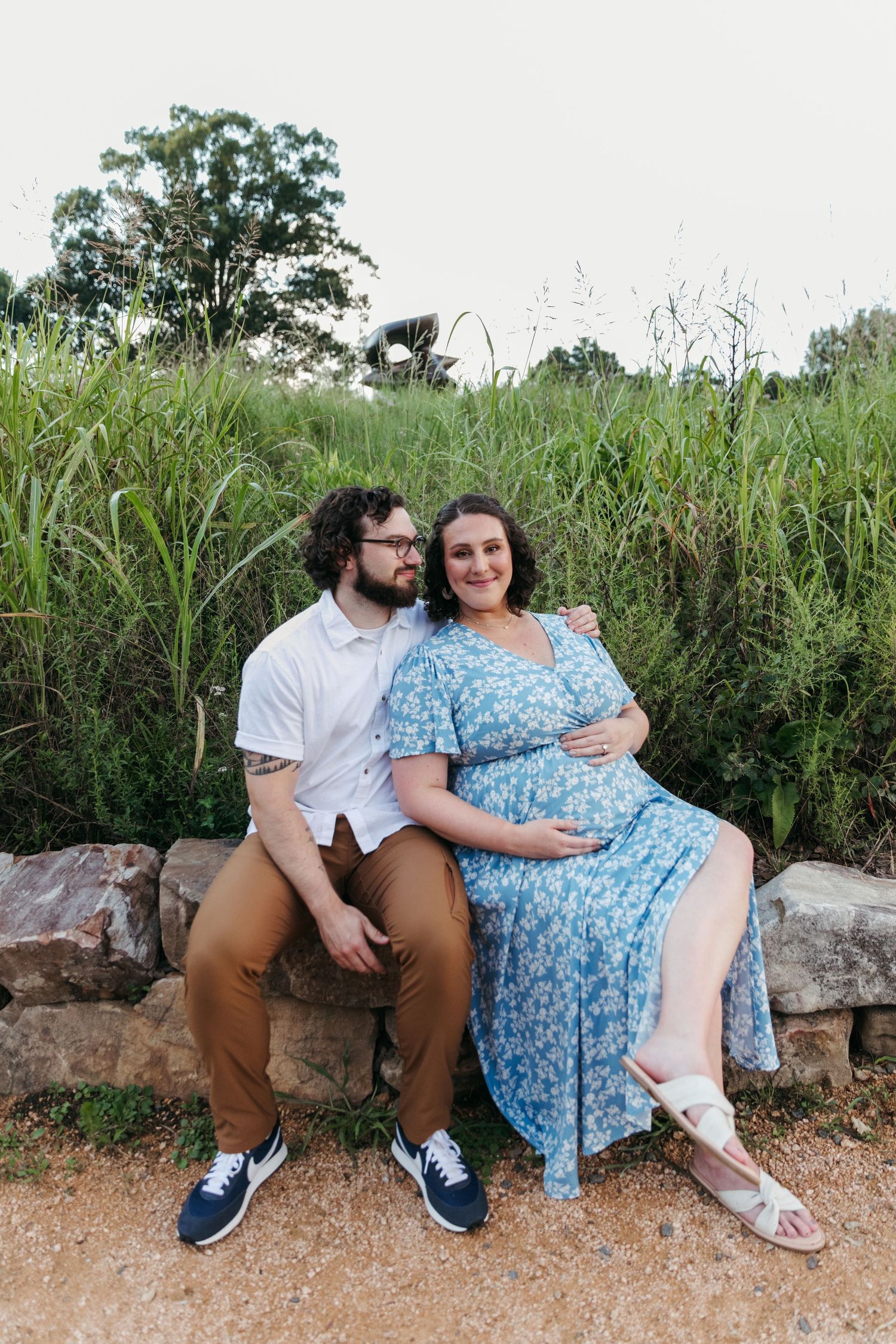 Two people sitting together on stone wall surrounded by tall grass and greenery.