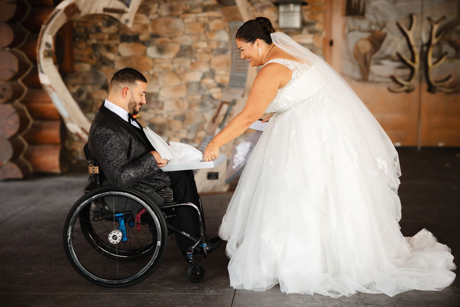 Bride in white tulle gown sharing a tender moment with groom in wheelchair indoors.