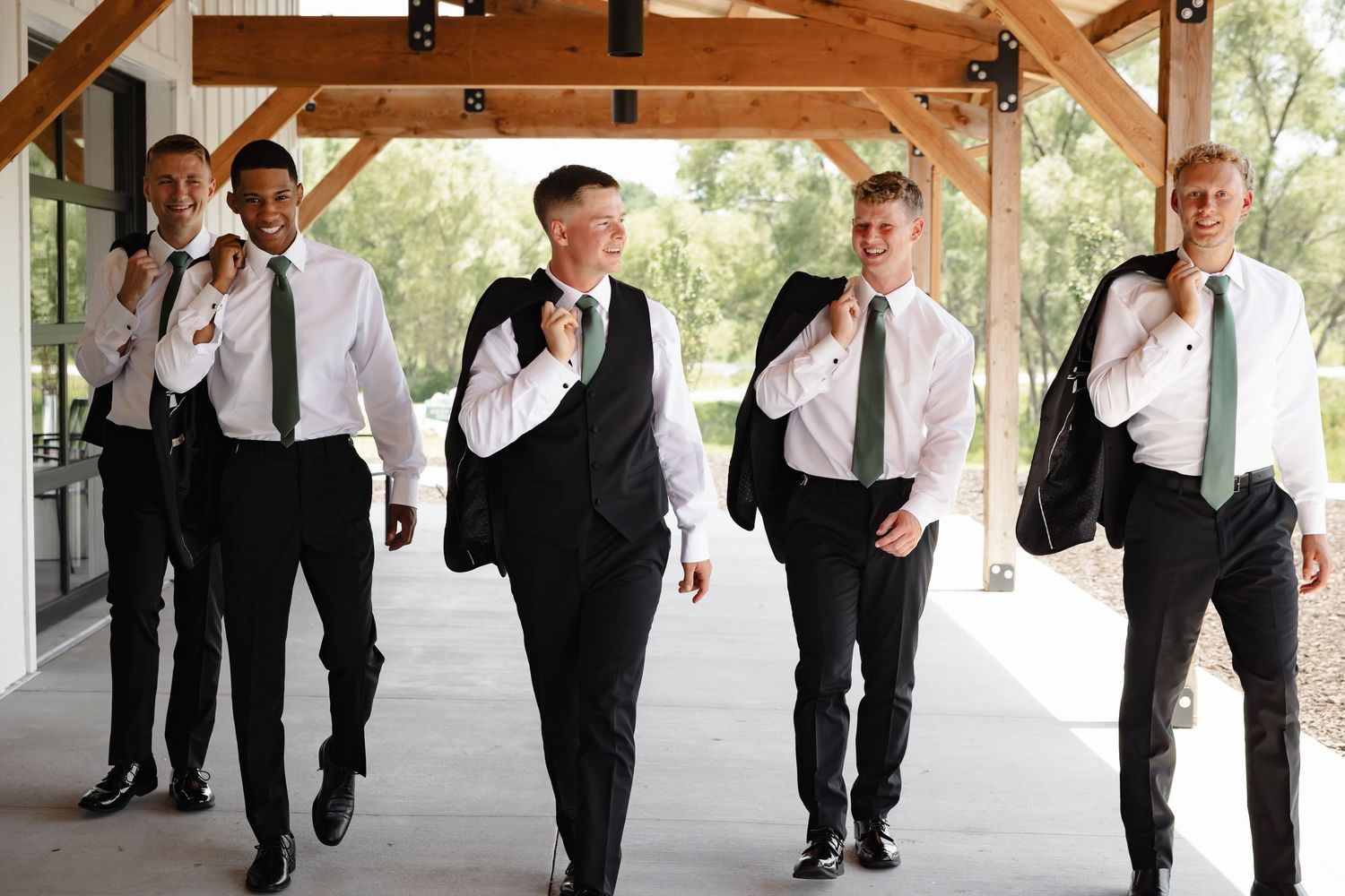 Five groomsmen in black suits and green ties walk confidently under a wooden pavilion at an outdoor wedding venue.
