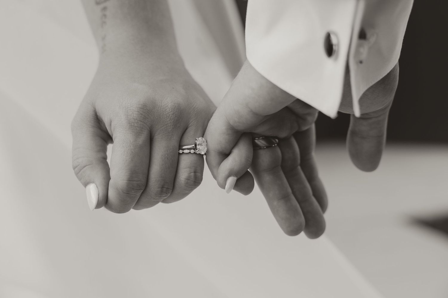 Black and white photo of a bride and groom holding pinkies, showcasing a diamond wedding ring set.
