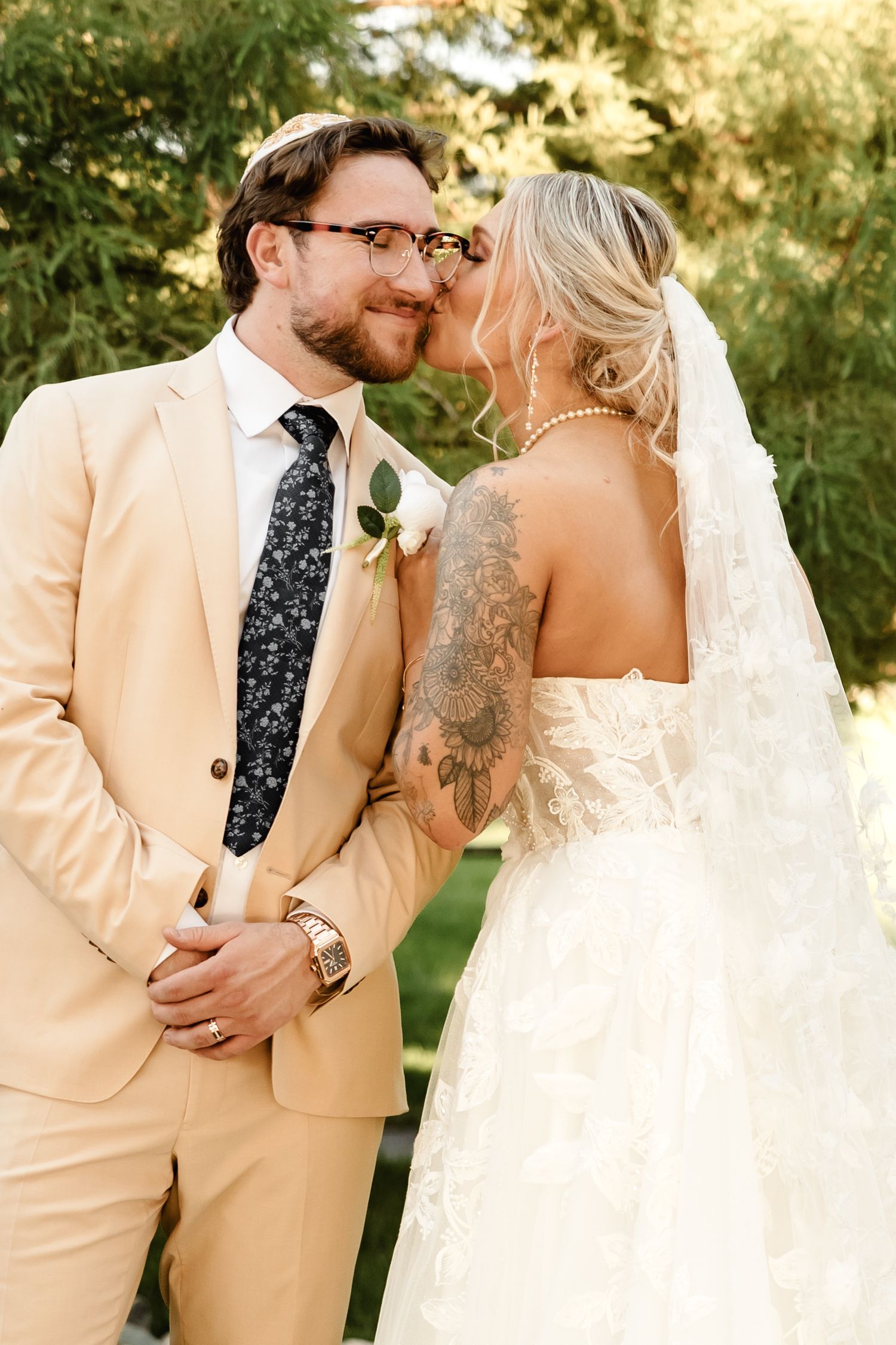A bride with a sunflower arm tattoo and veil kisses a groom in a tan suit with floral tie outdoors.