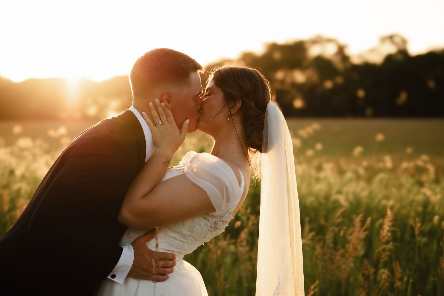 A bride and groom share a romantic kiss in a golden sunlit field during their outdoor wedding ceremony.