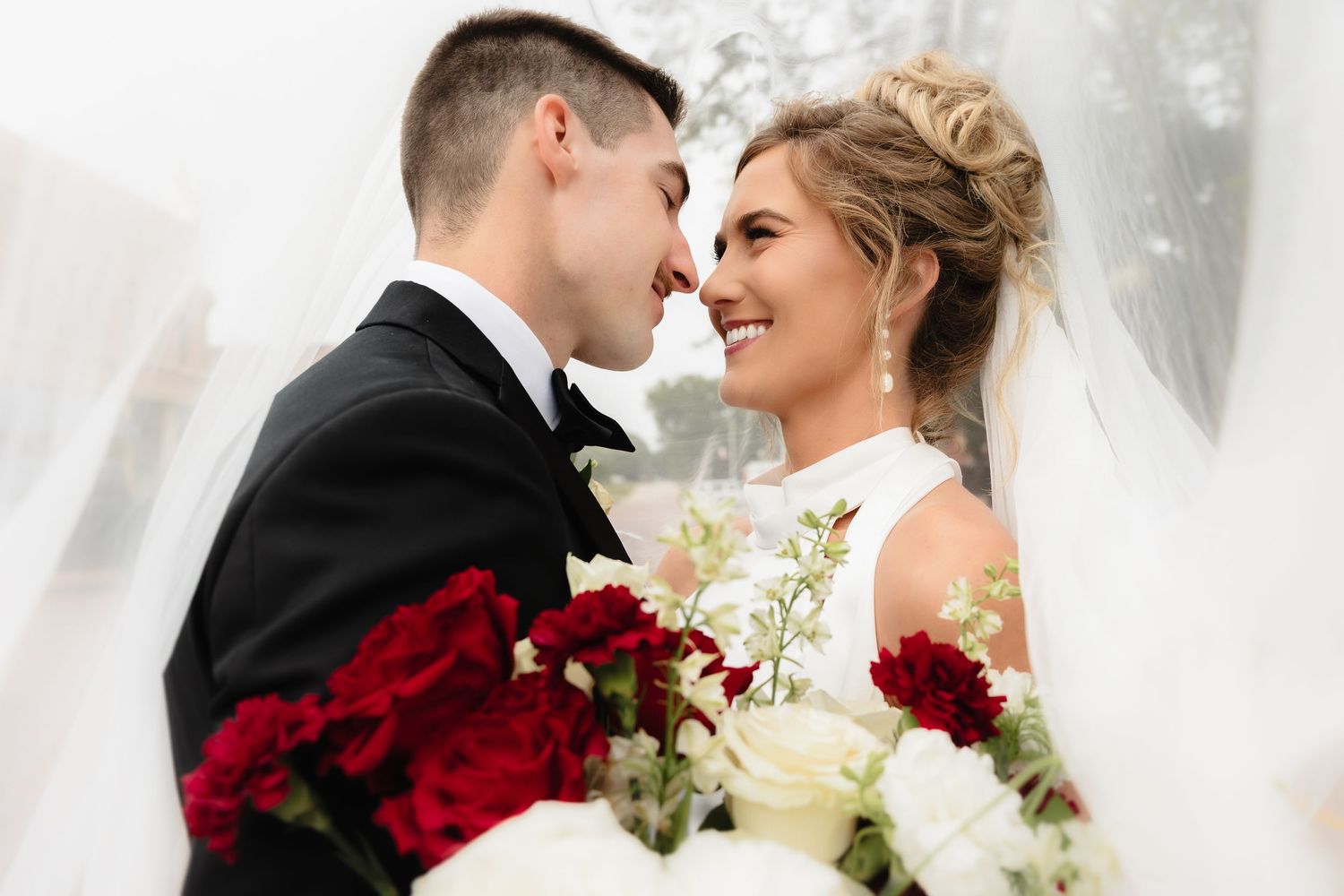 Bride and groom smile under veil with red and white rose bouquet.