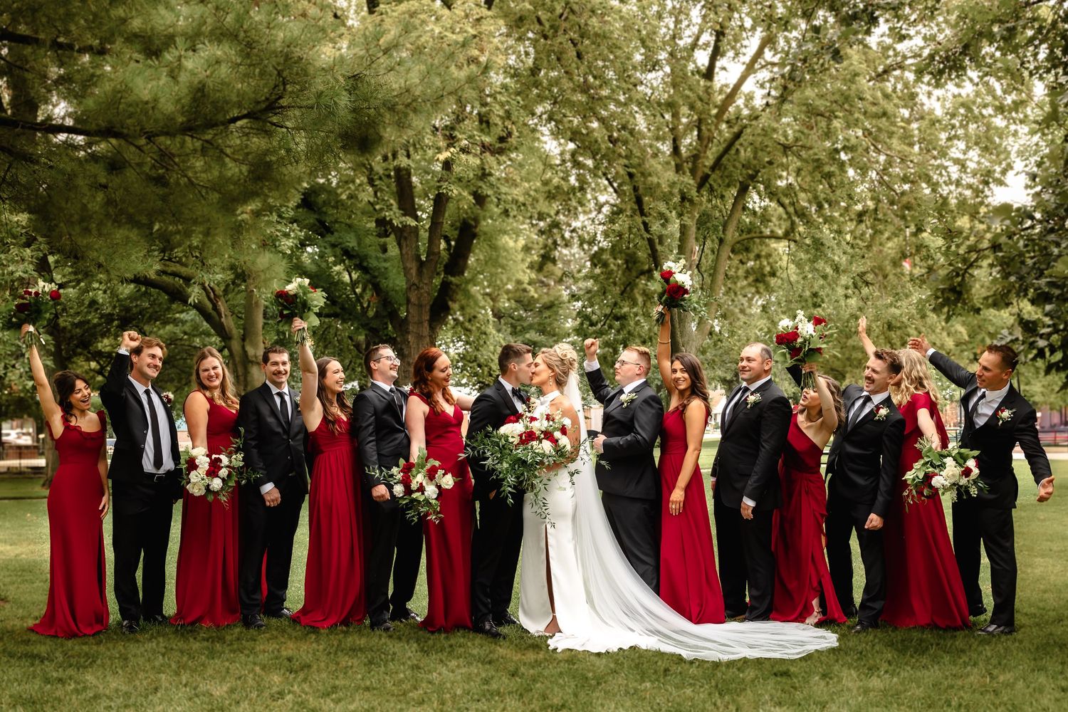 Wedding party in red dresses and black suits celebrating under green trees.