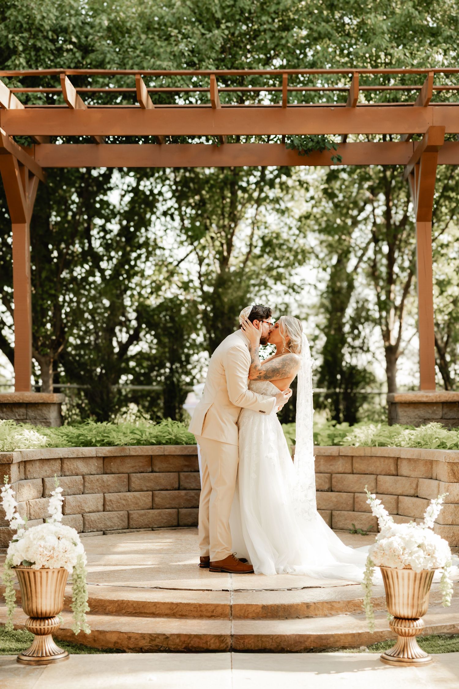 A bride and groom share their first kiss under a wooden pergola surrounded by lush greenery and white floral arrangements.