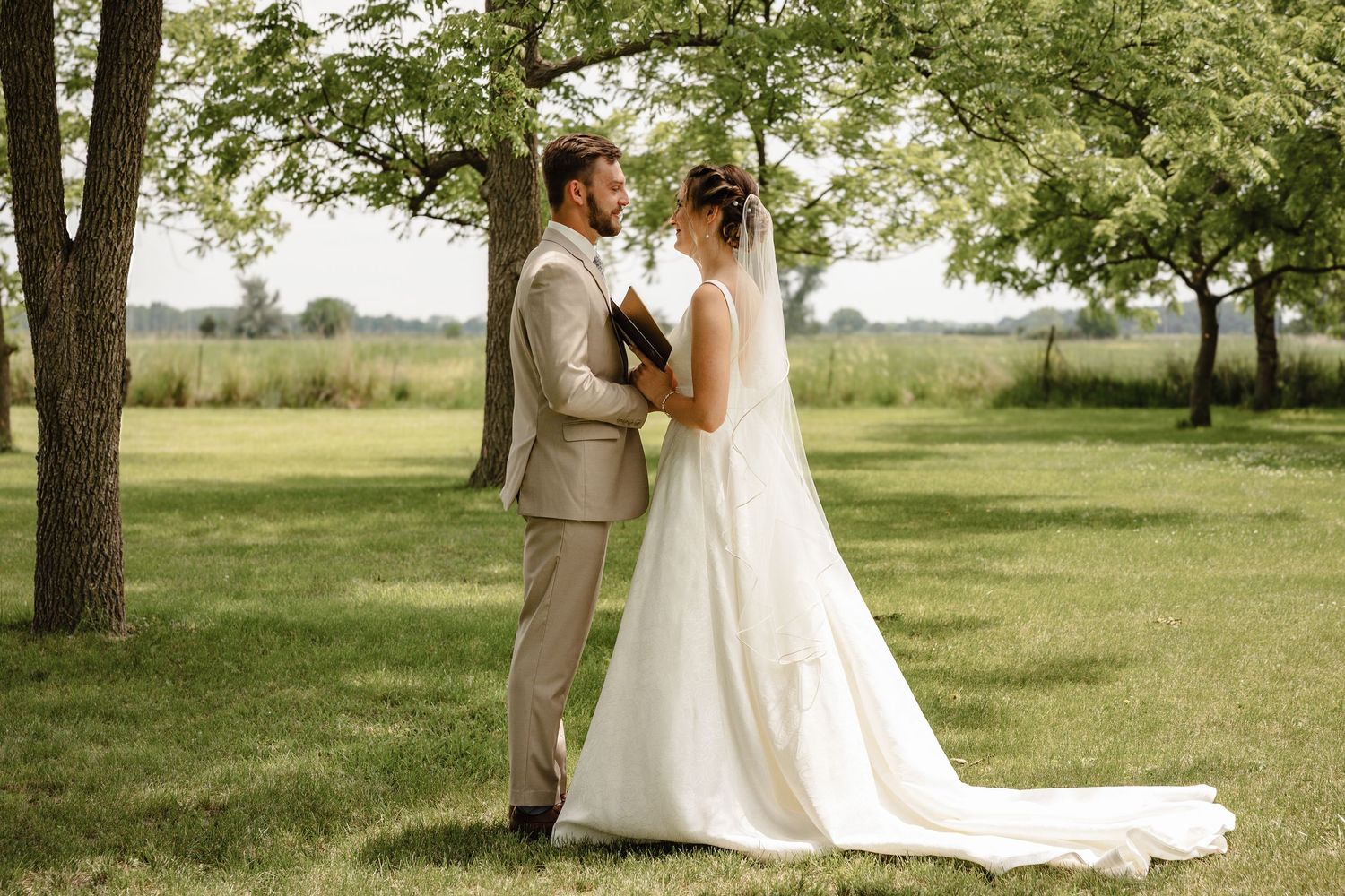 Bride and groom share a tender moment standing together under green trees in an outdoor wedding setting.