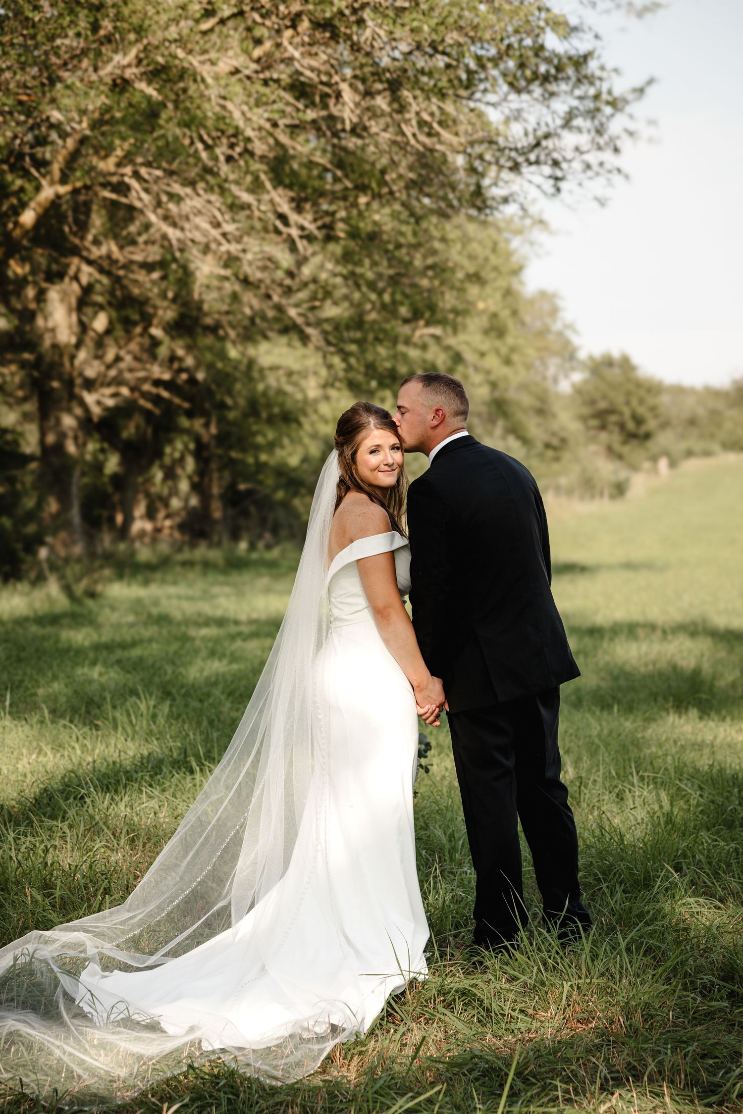 Bride in white off-shoulder gown with long veil holds hands with groom in black suit outdoors in green field.