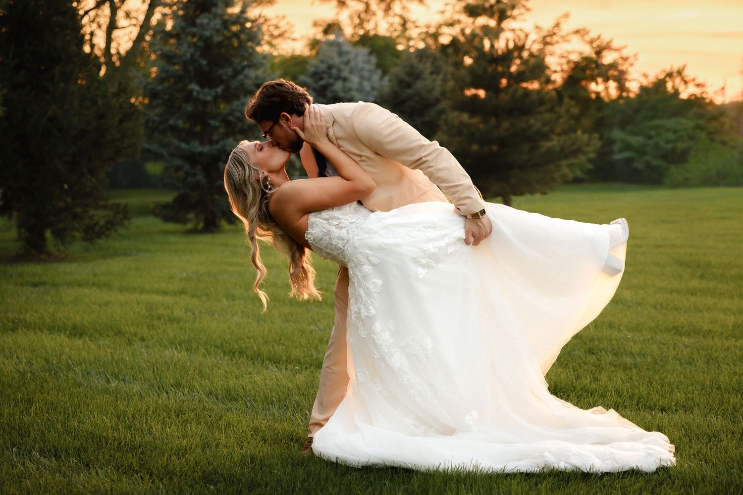 Groom dips bride for a romantic kiss at golden hour on a lush green lawn.
