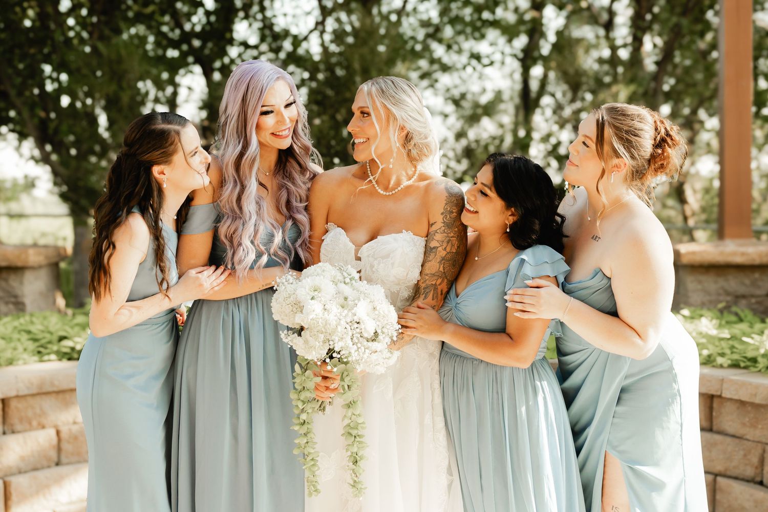 Bride with bouquet laughing with bridesmaids in sage green dresses outdoors.