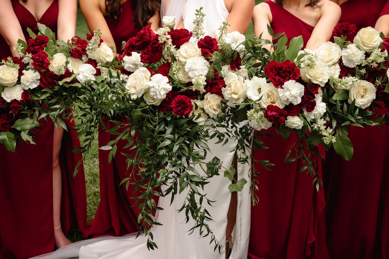 Bridesmaids in red gowns holding lush bouquets of red and white roses with greenery at a wedding.