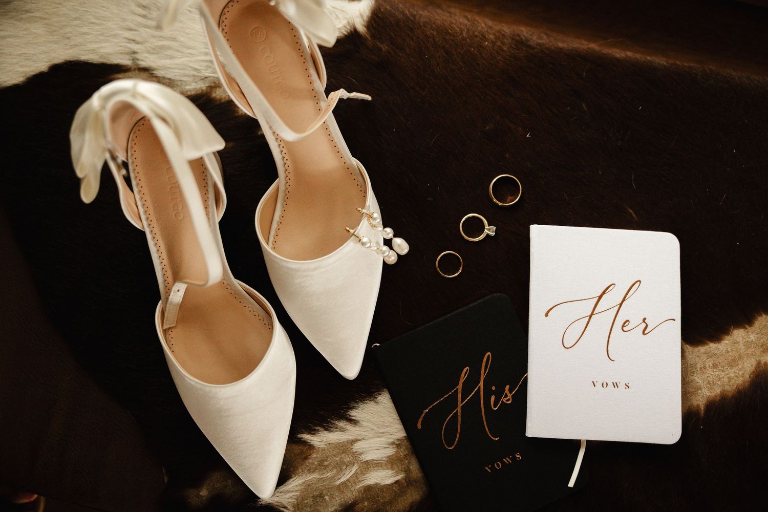 Flat lay of white bridal heels, gold rings, and His and Her vow books.