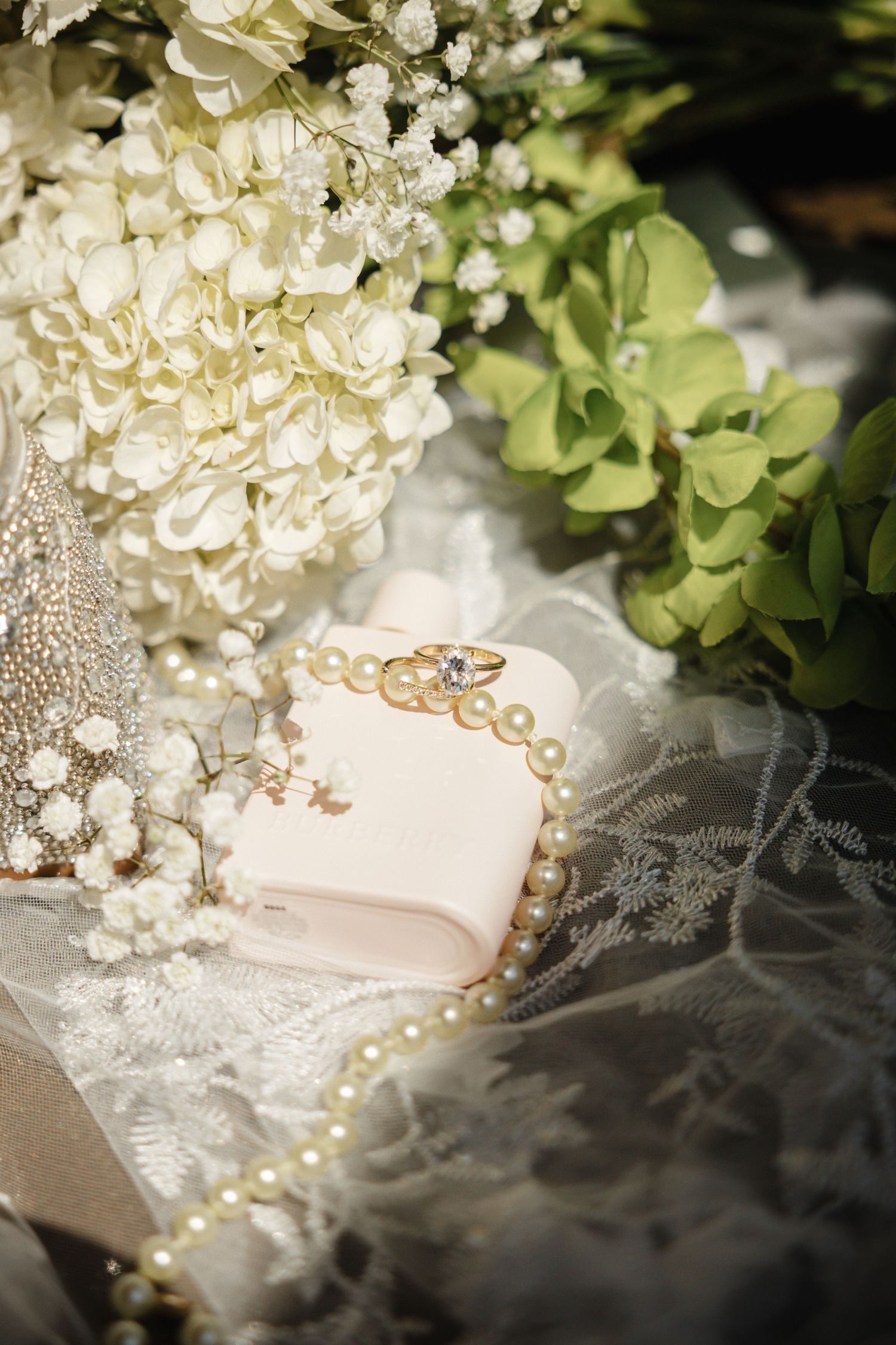 Elegant wedding flat lay with diamond engagement ring on pearl bracelet, white hydrangeas, baby's breath, and lace veil.
