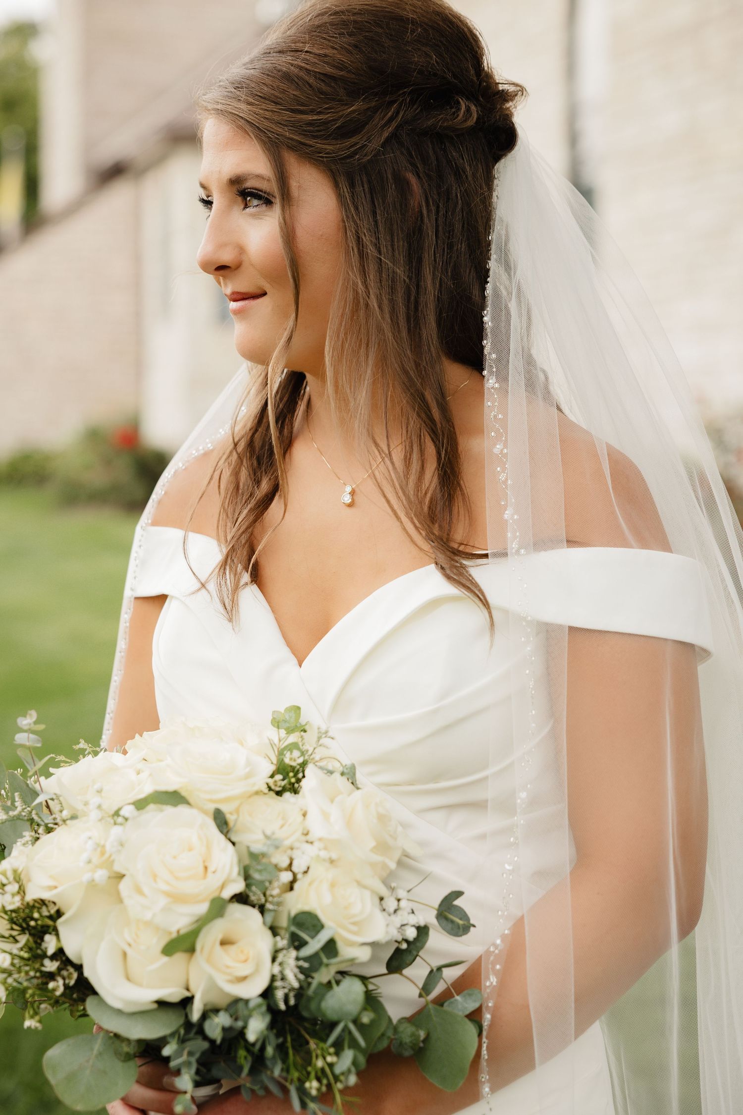 Bride in off-shoulder gown holding white rose bouquet with veil outdoors.