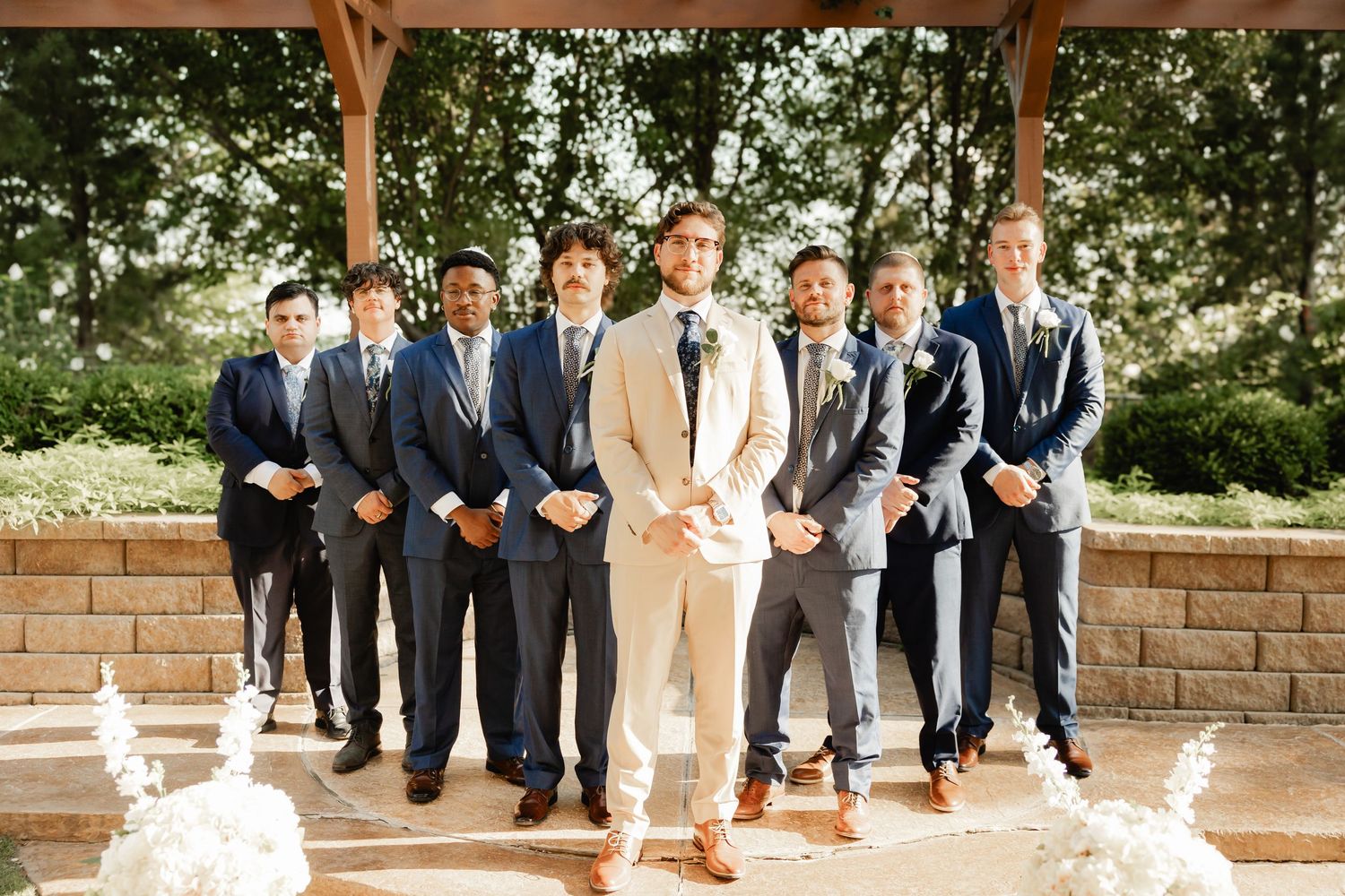 Groom in tan suit poses with groomsmen in navy and gray suits under a pavilion.