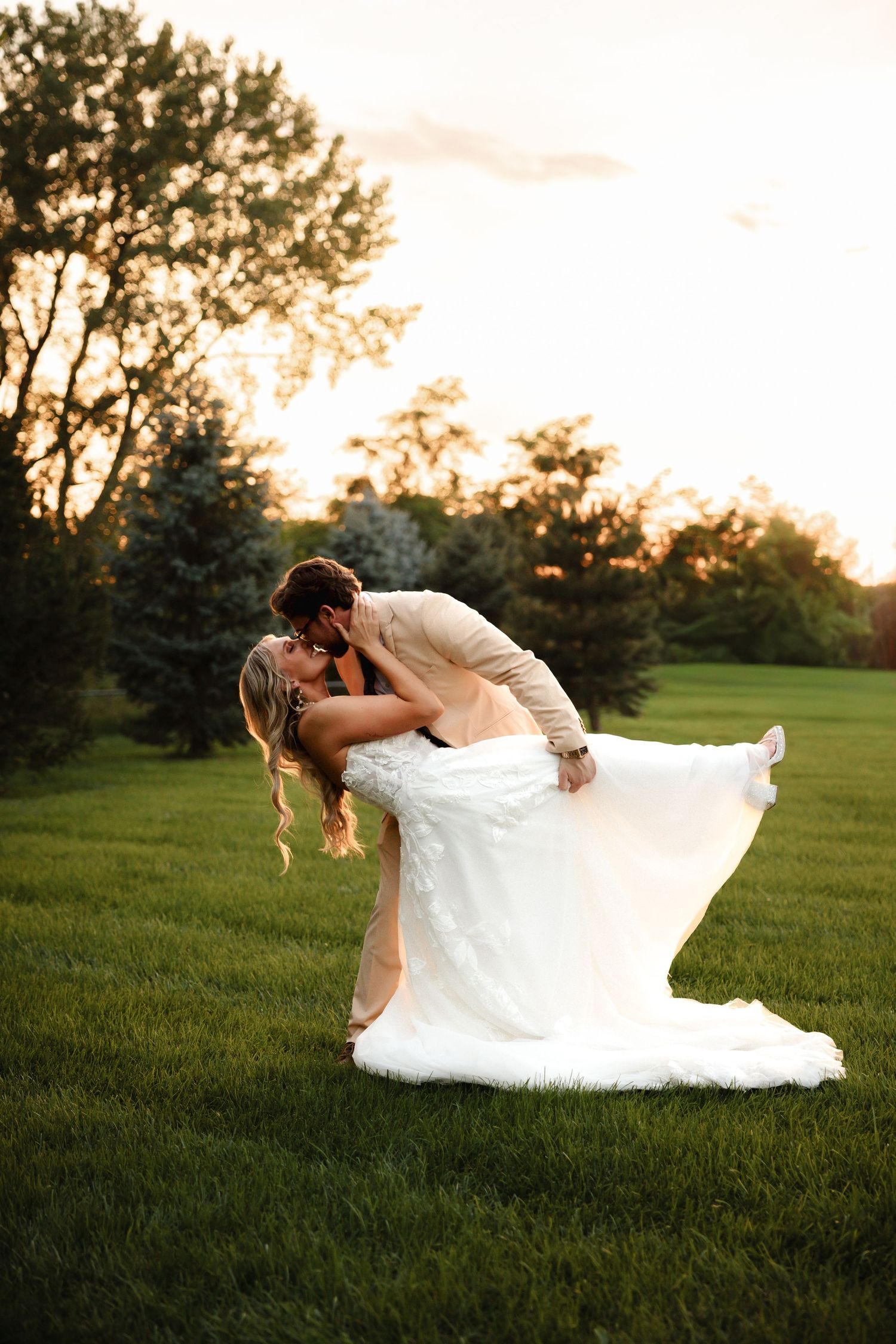 Groom dips bride in white wedding gown at sunset on green lawn.