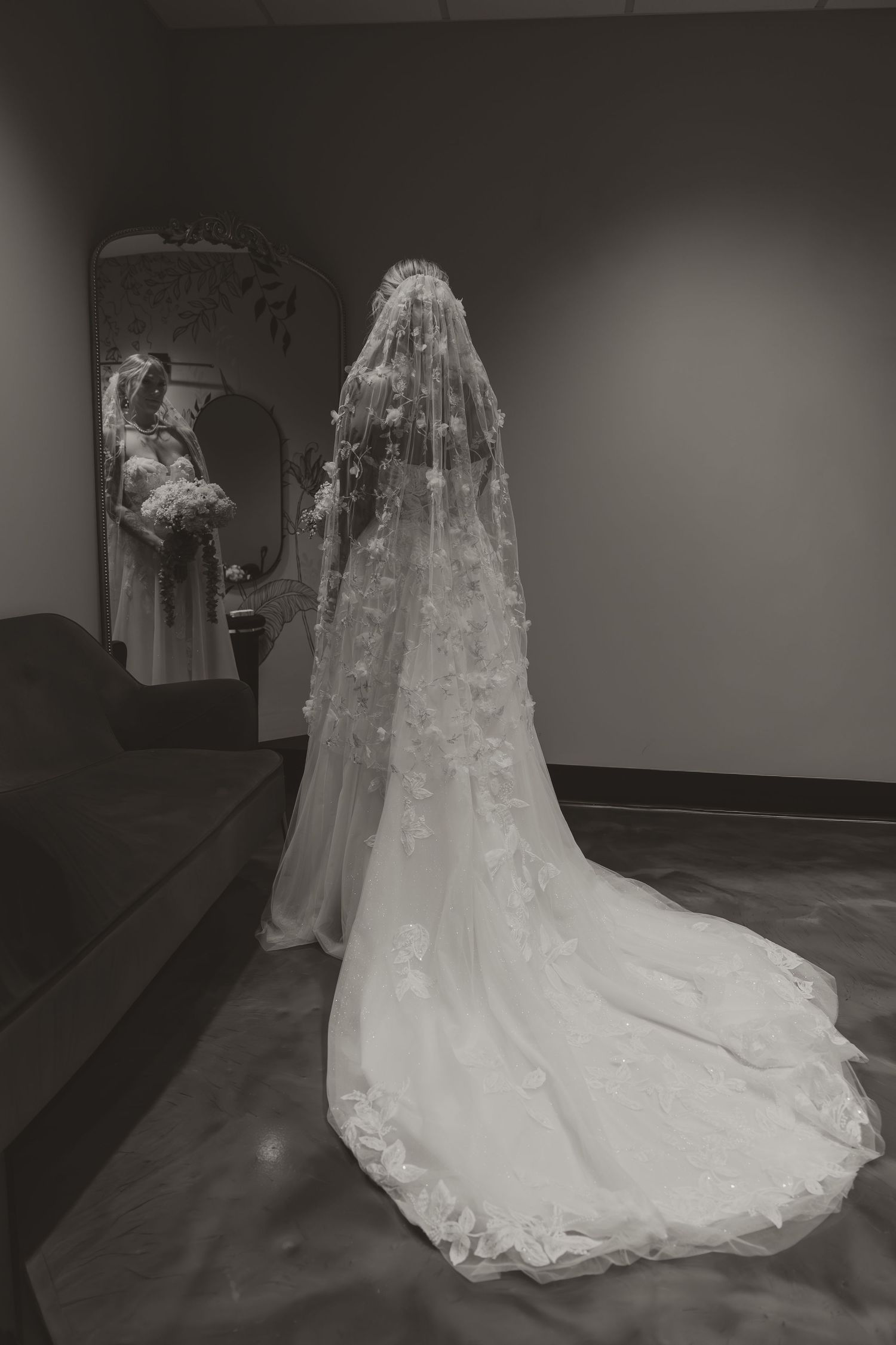 Black and white photo of a bride in a lace wedding gown and cathedral veil, viewed from behind near a mirror.