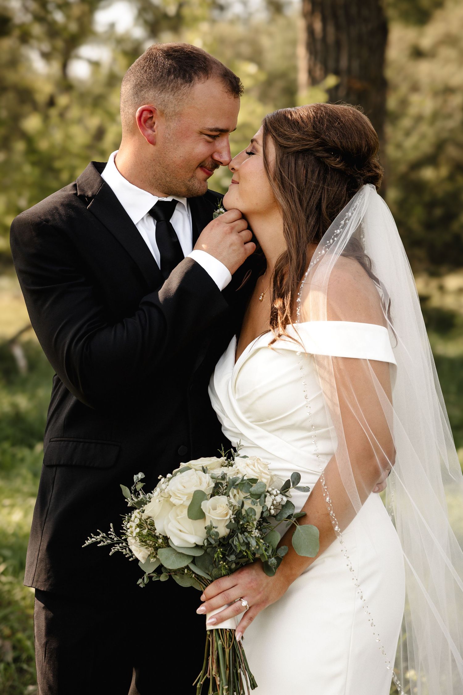 A bride and groom share a romantic moment outdoors, she holds a white bouquet while wearing a veil and off-shoulder gown.