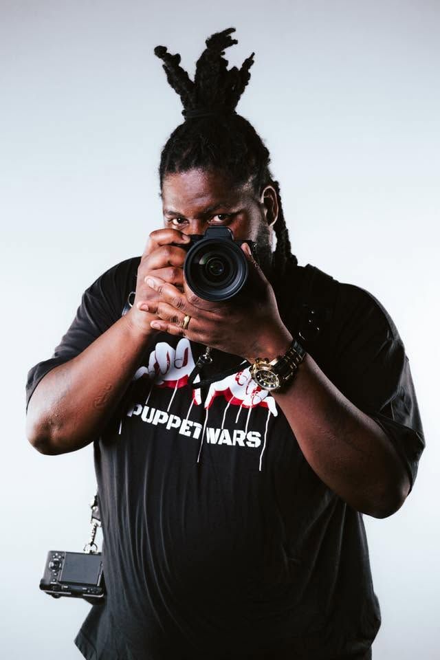 Photographer with dreadlocks aims DSLR camera wearing Puppet Wars black tee with gold accessories.