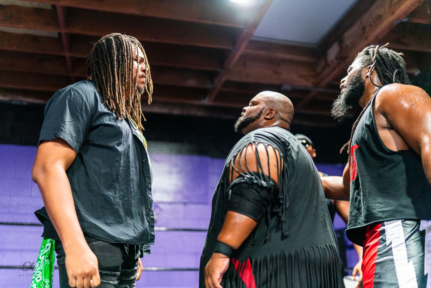 Three wrestlers in black attire face off in dramatic confrontation inside a wrestling arena.