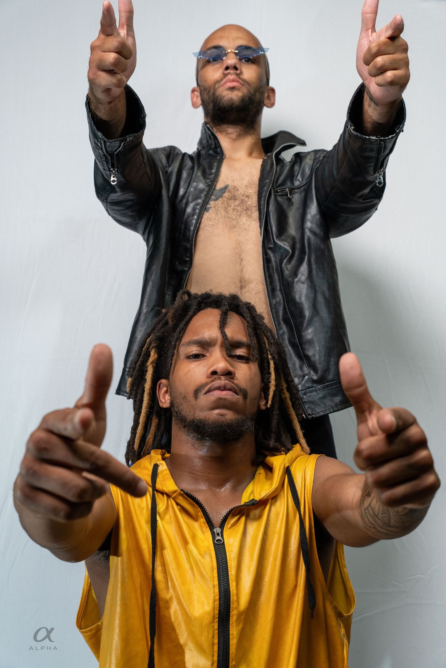 Two men pose confidently, one in yellow vest with dreadlocks pointing forward, another in open leather jacket gesturing upward.