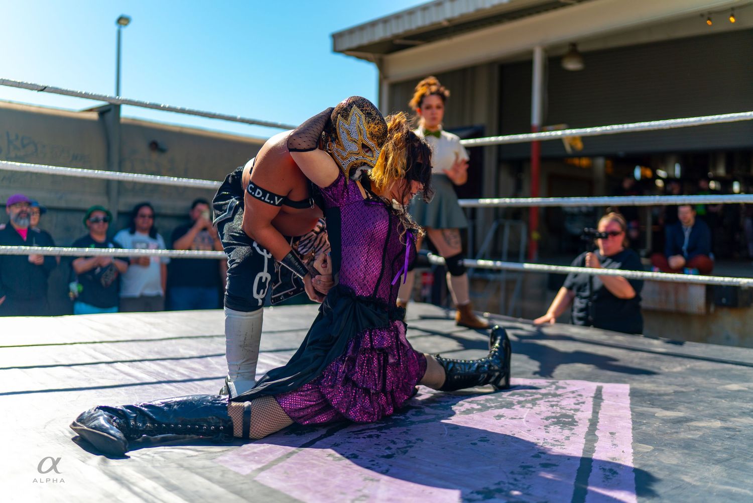 Two wrestlers grapple on outdoor ring mat, one in colorful costume holds opponent down during match.