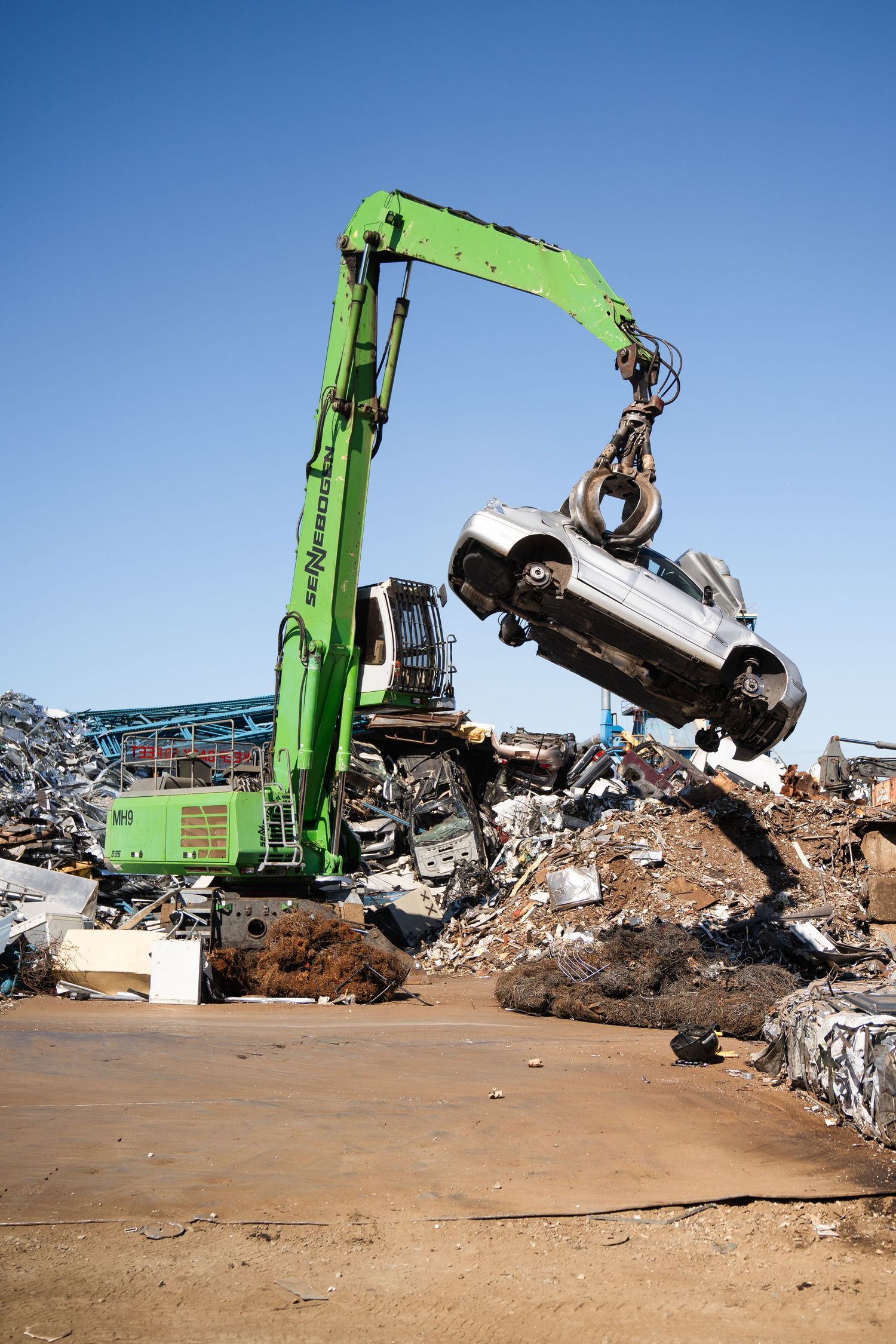 Green SENNEBOGEN material handler lifting a crushed car at an outdoor scrap metal recycling yard.