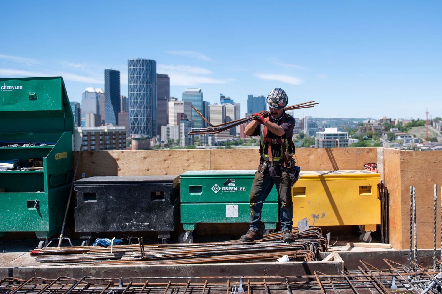 Construction worker in safety gear carrying materials on a rooftop with a city skyline in the background.