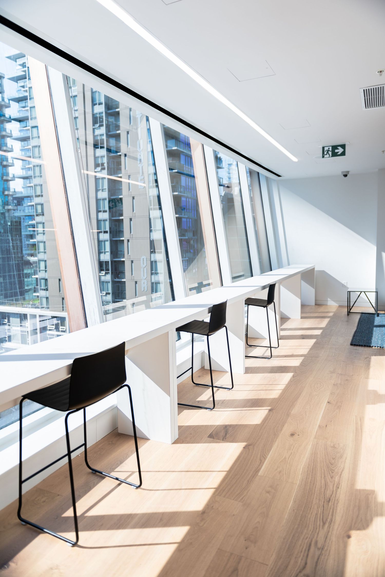 Bright modern workspace with a long white counter, black bar stools, and floor-to-ceiling windows.