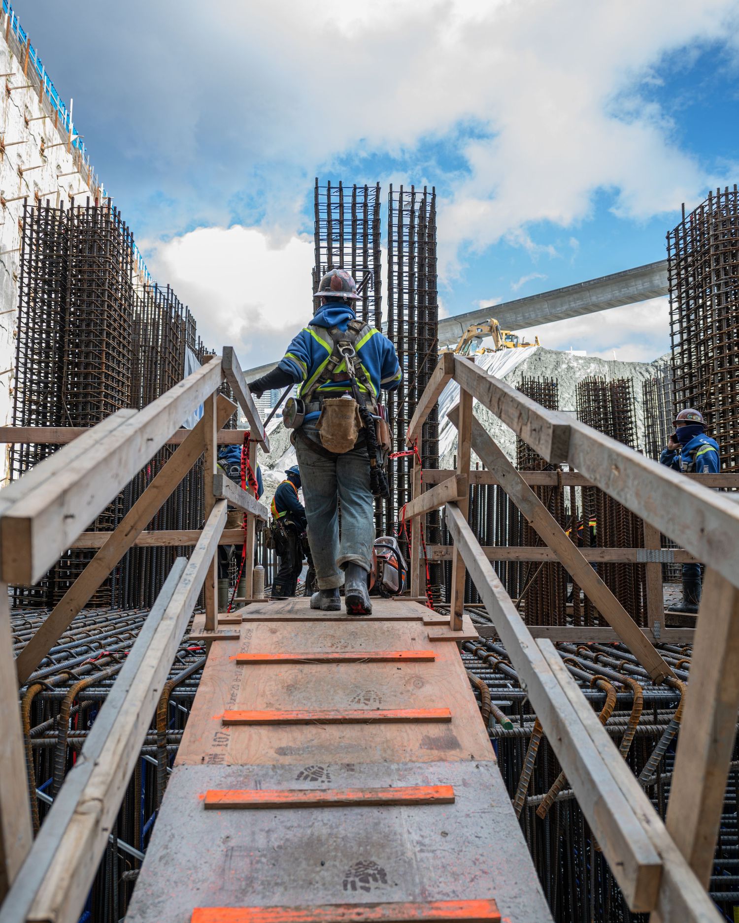 Construction worker in safety gear climbing a ramp surrounded by rebar columns at a building site.