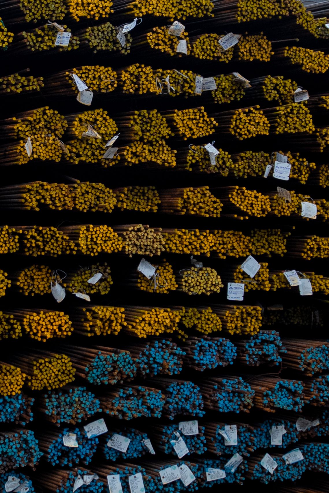 Stacked bundles of yellow and blue color-coded steel rebar rods with identification tags in a warehouse.