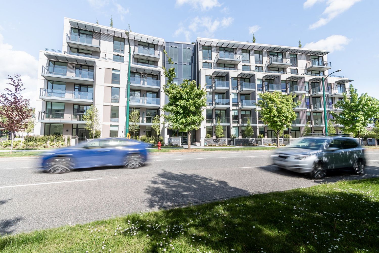 Contemporary mid-rise residential building with balconies viewed from across a busy street on a sunny day.