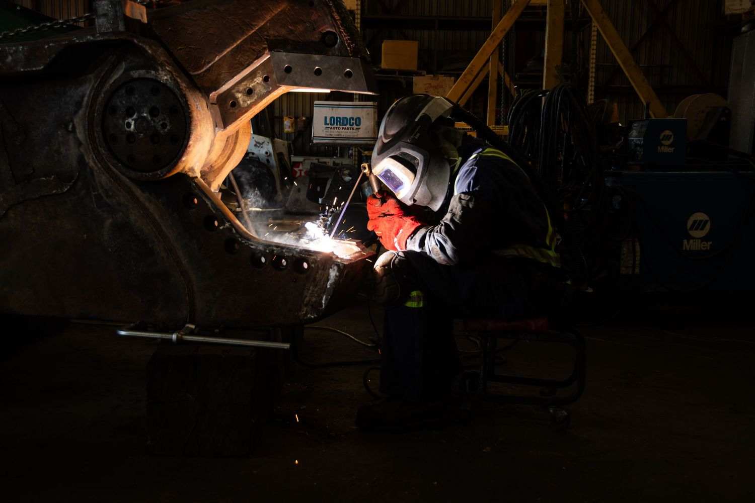 Welder in protective gear creating bright sparks while welding heavy industrial machinery in dark workshop.