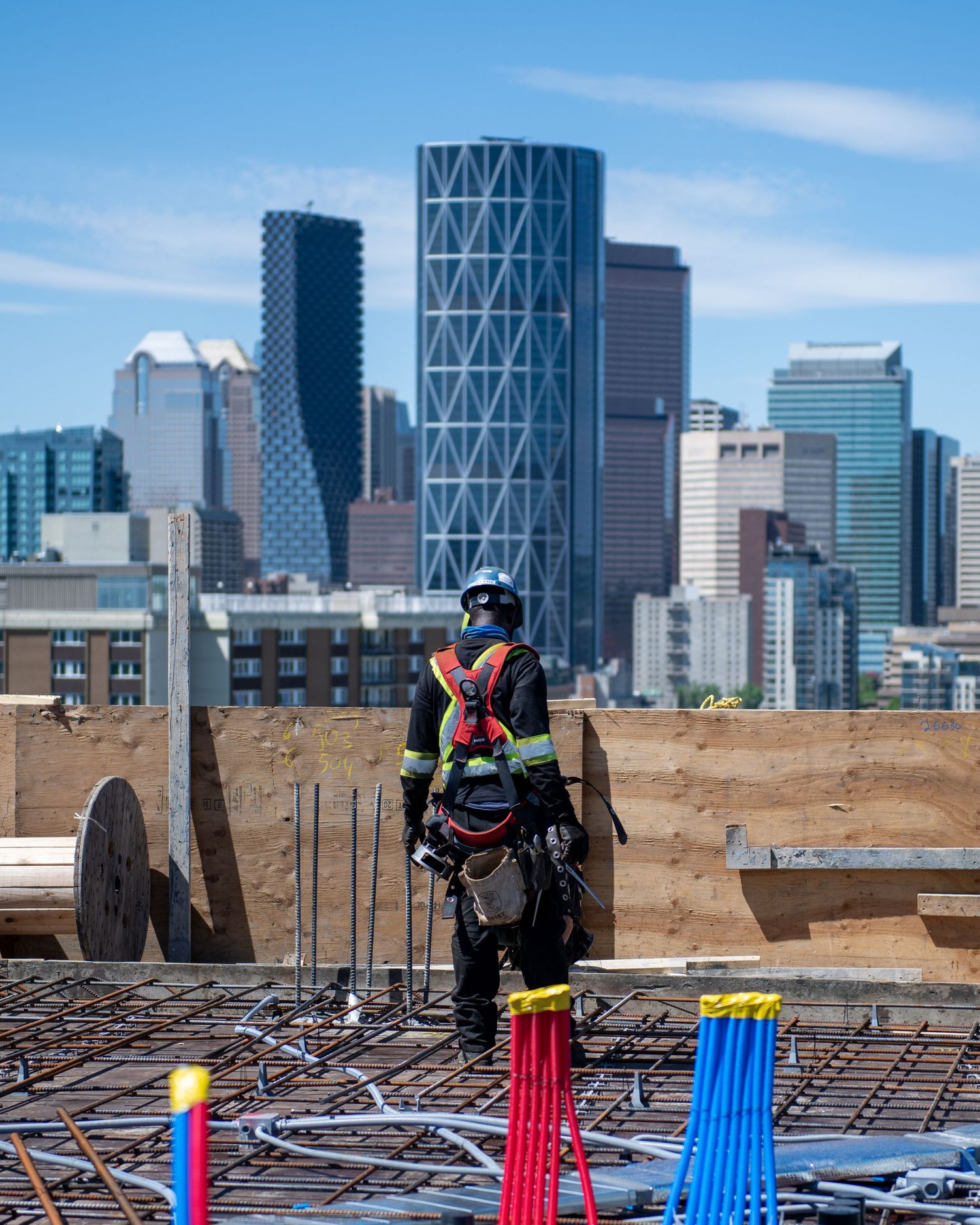 Construction worker in safety harness standing on rebar-reinforced concrete slab with downtown city skyline behind.