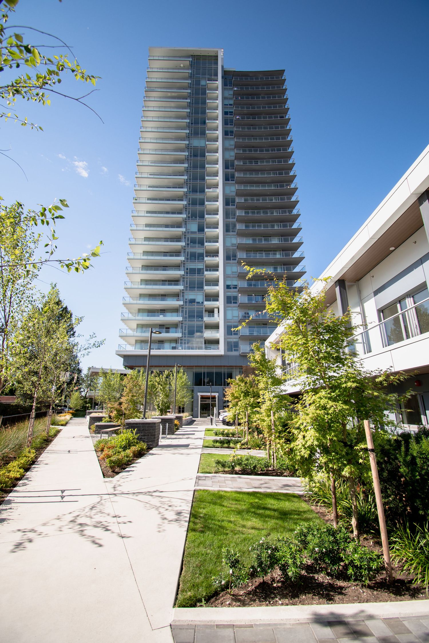 Modern residential high-rise tower with landscaped pathways and green trees on a clear sunny day.