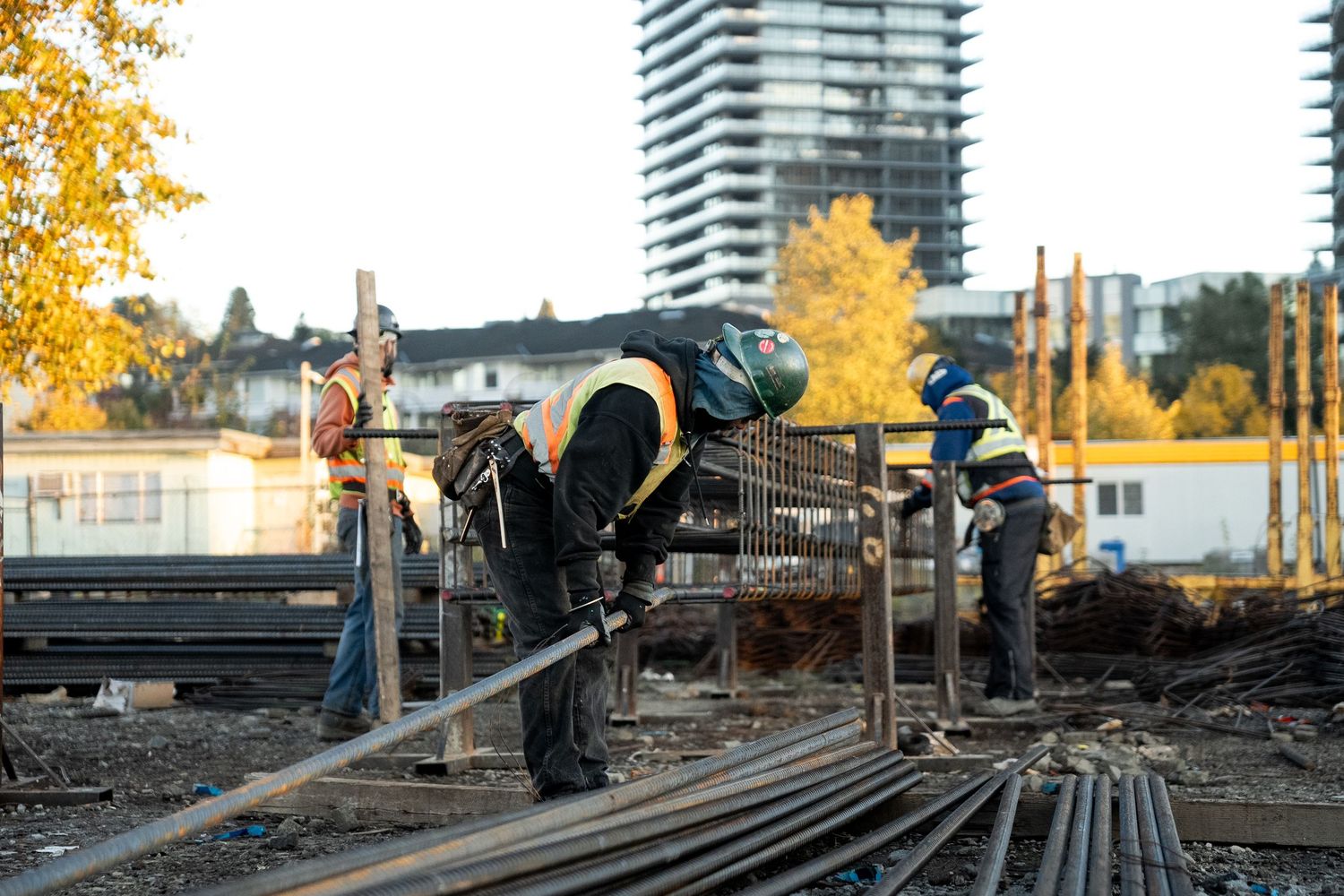 Two construction workers bending and cutting steel rebar at an outdoor job site in autumn.
