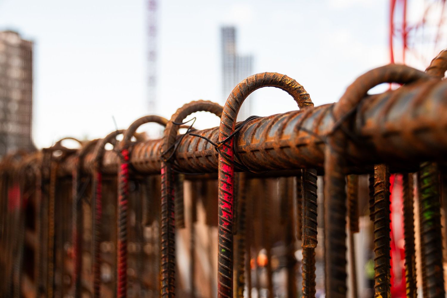 Close-up of rusty rebar loops at a construction site with city buildings in the blurred background.