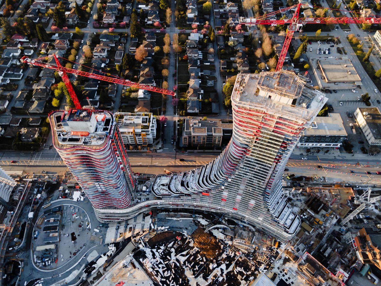 Aerial view of two curved high-rise towers under construction with red cranes in urban neighborhood.