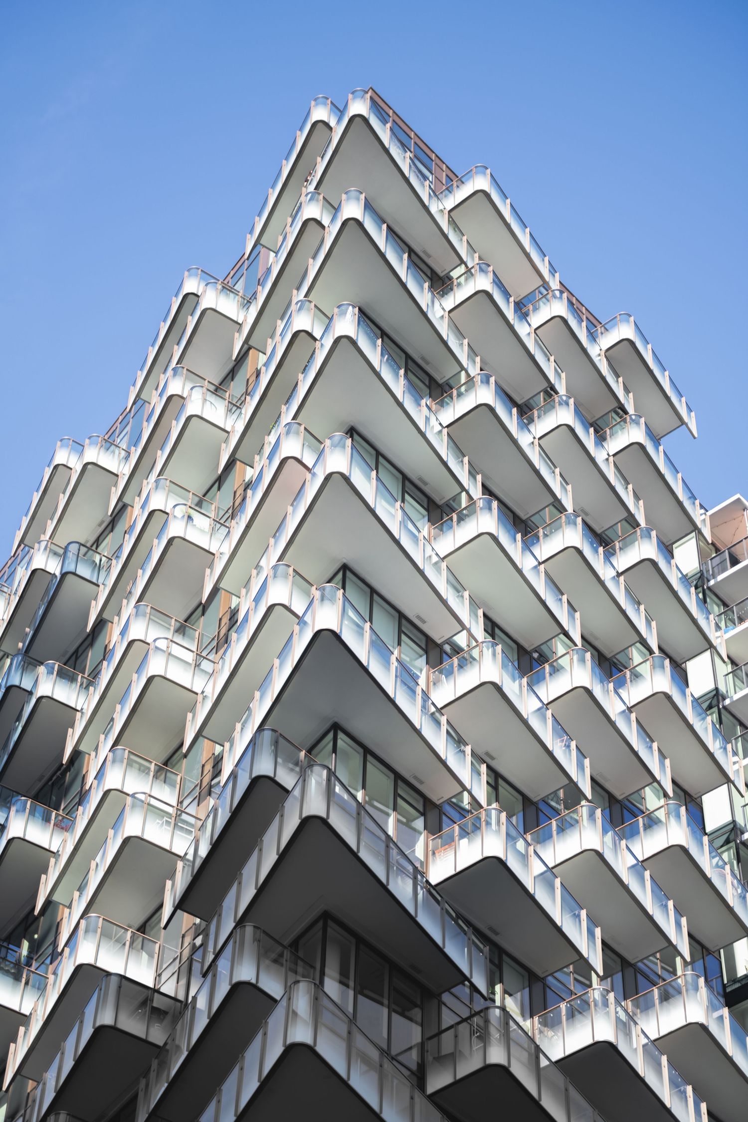Low angle view of contemporary white residential apartment building with geometric staggered balconies against blue sky.